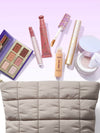 tarte Collection of makeup products including lipsticks, a palette, and a cream on a light purple background.
