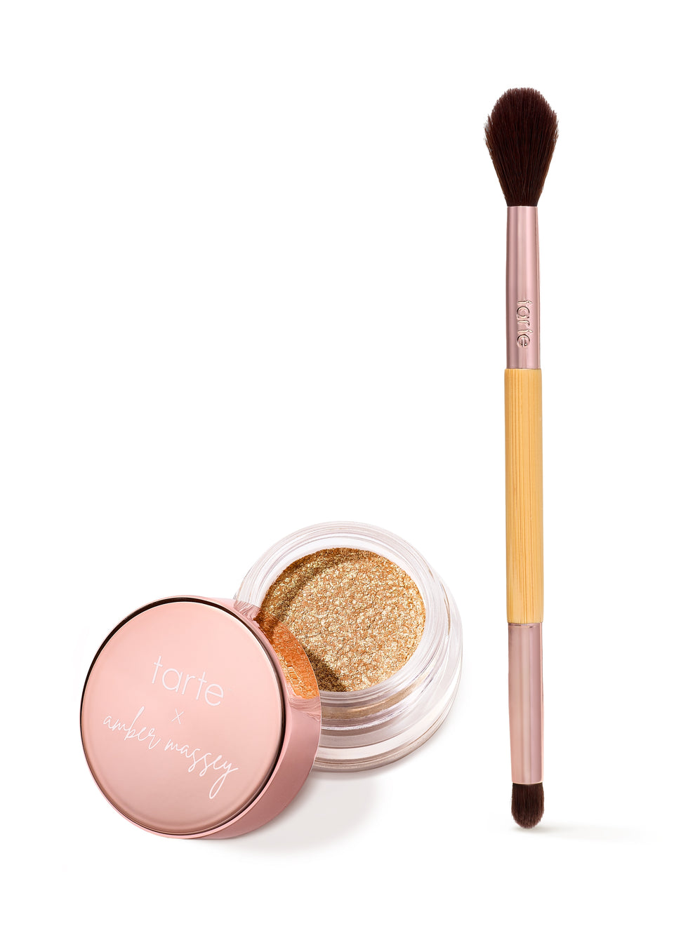 An open jar of shimmery gold eyeshadow sits alongside a dual-ended makeup brush; the lid reads tarte x amber massey. The background is plain white, emphasizing the products.
