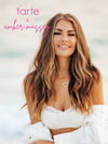 A woman with long, wavy hair smiles, wearing a white top and heart necklace, standing in a serene, beach-like setting. Visible text: tarte™ x amber massey.