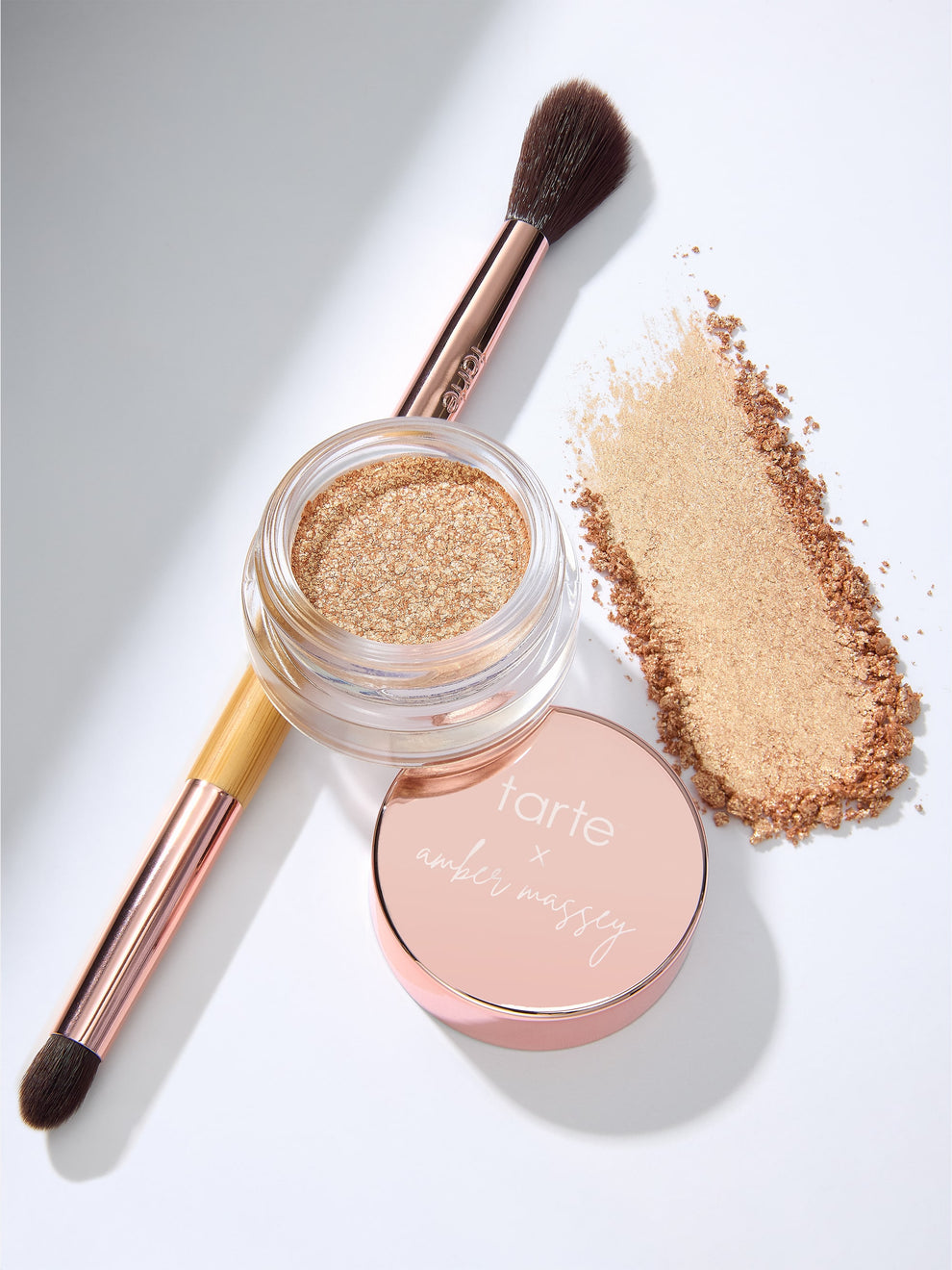 An open jar of shimmery eyeshadow sits beside a makeup brush and its scattered powder. The lid, bearing tarte x amber massey, rests nearby on a light background.
