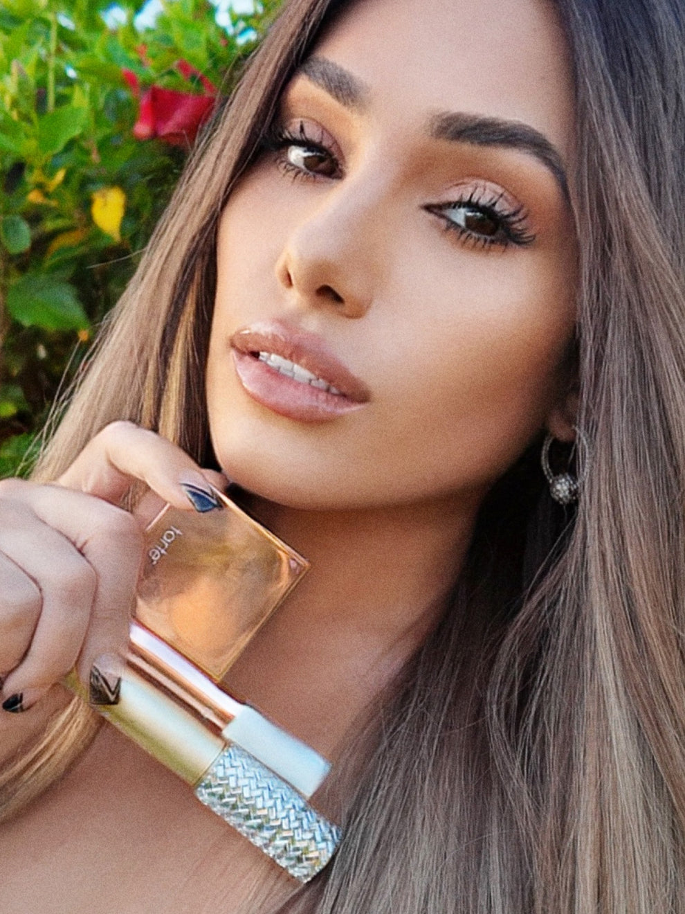 A person holds Tarte Cosmetics products, including a compact and a tube, in a garden setting. They have long hair and wear earrings, with a red flower visible in the background.
