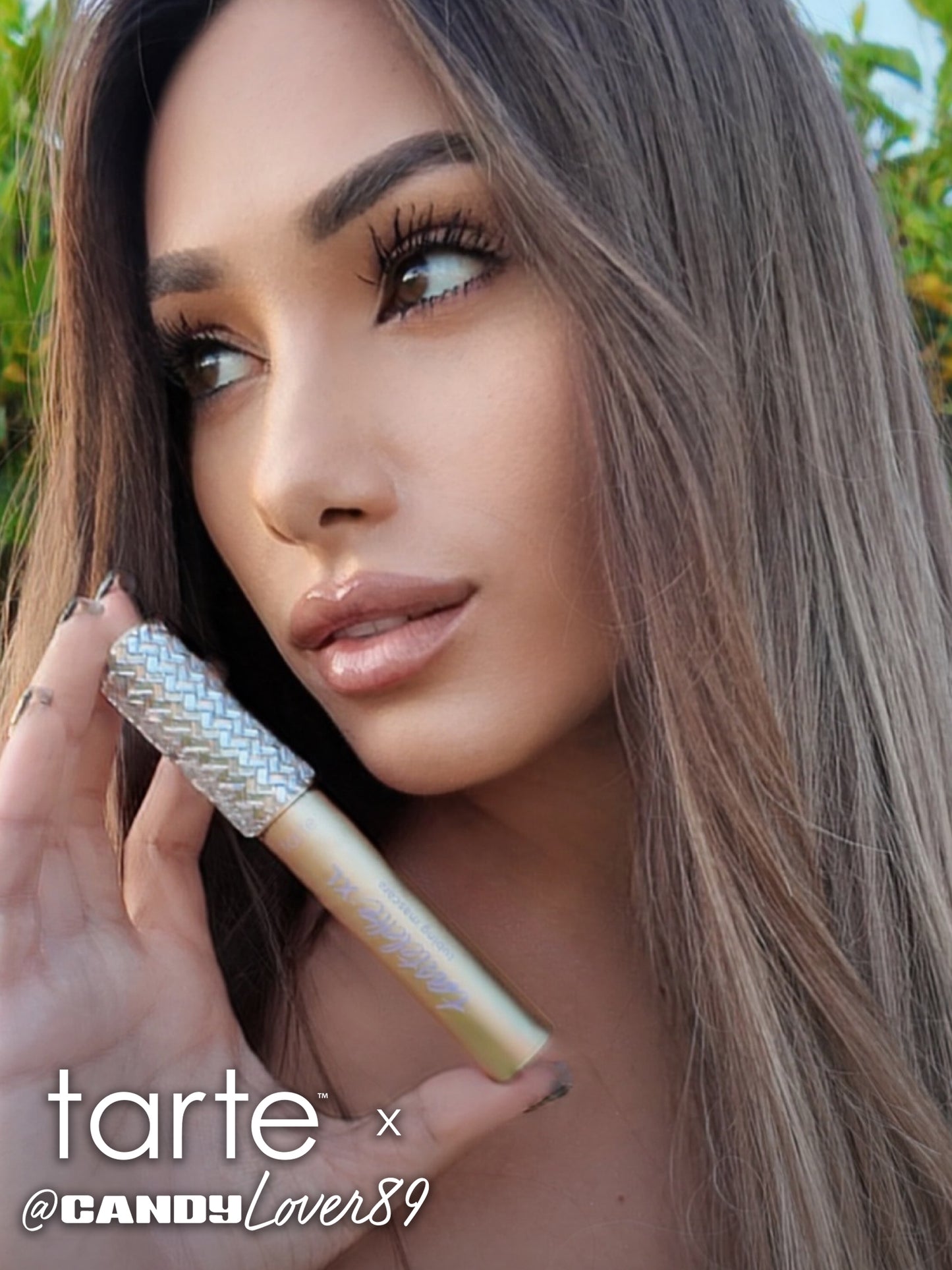 A person holds a shimmering gold Tarte Cosmetics mascara near their face, gazing sideways. Lush greenery forms the background. Text: tarte™ x @CANDYLover89.