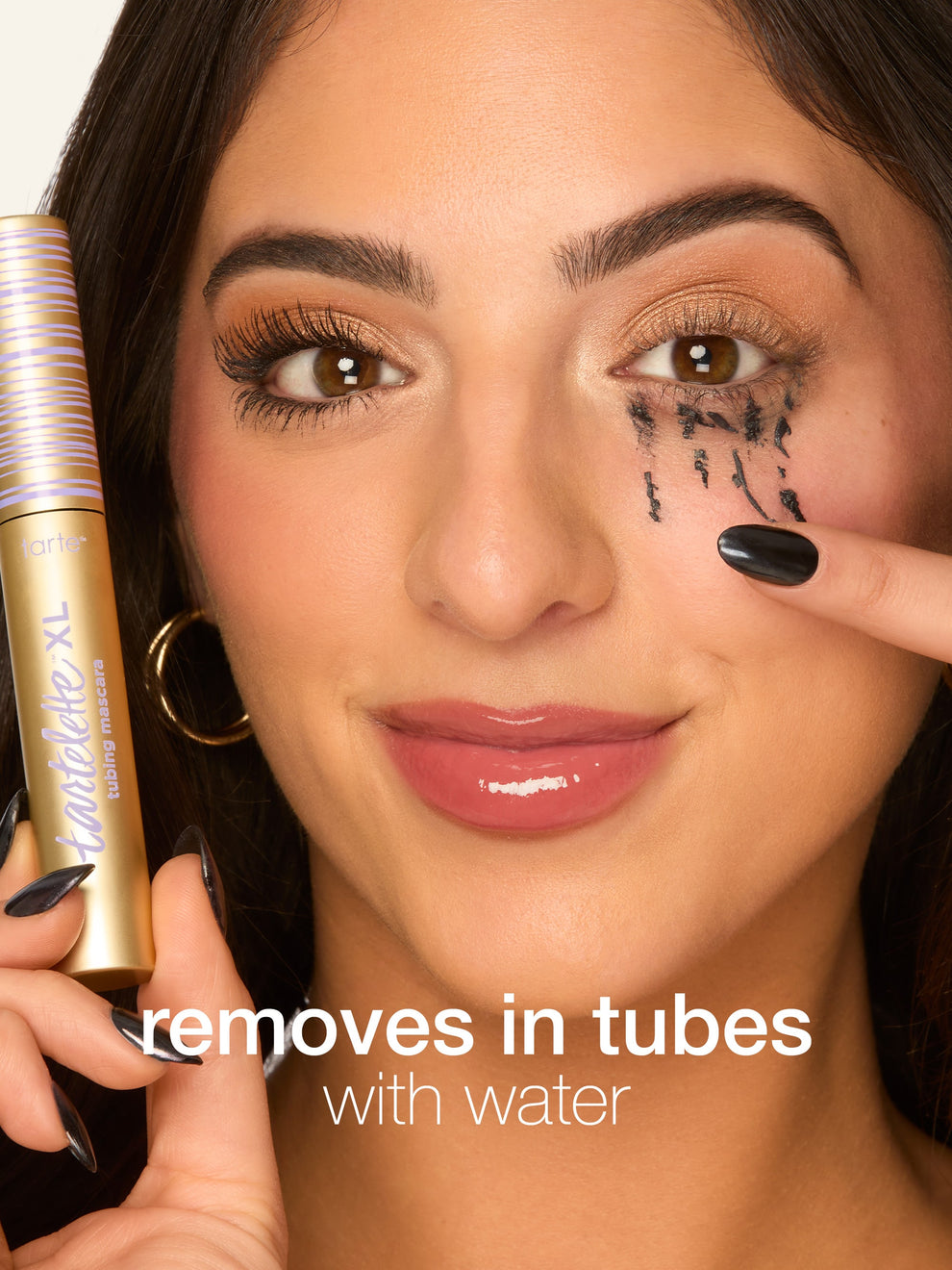 A person holds a Tarte Cosmetics tube mascara while gently removing tube-like makeup from their eye. Text reads, removes in tubes with water.