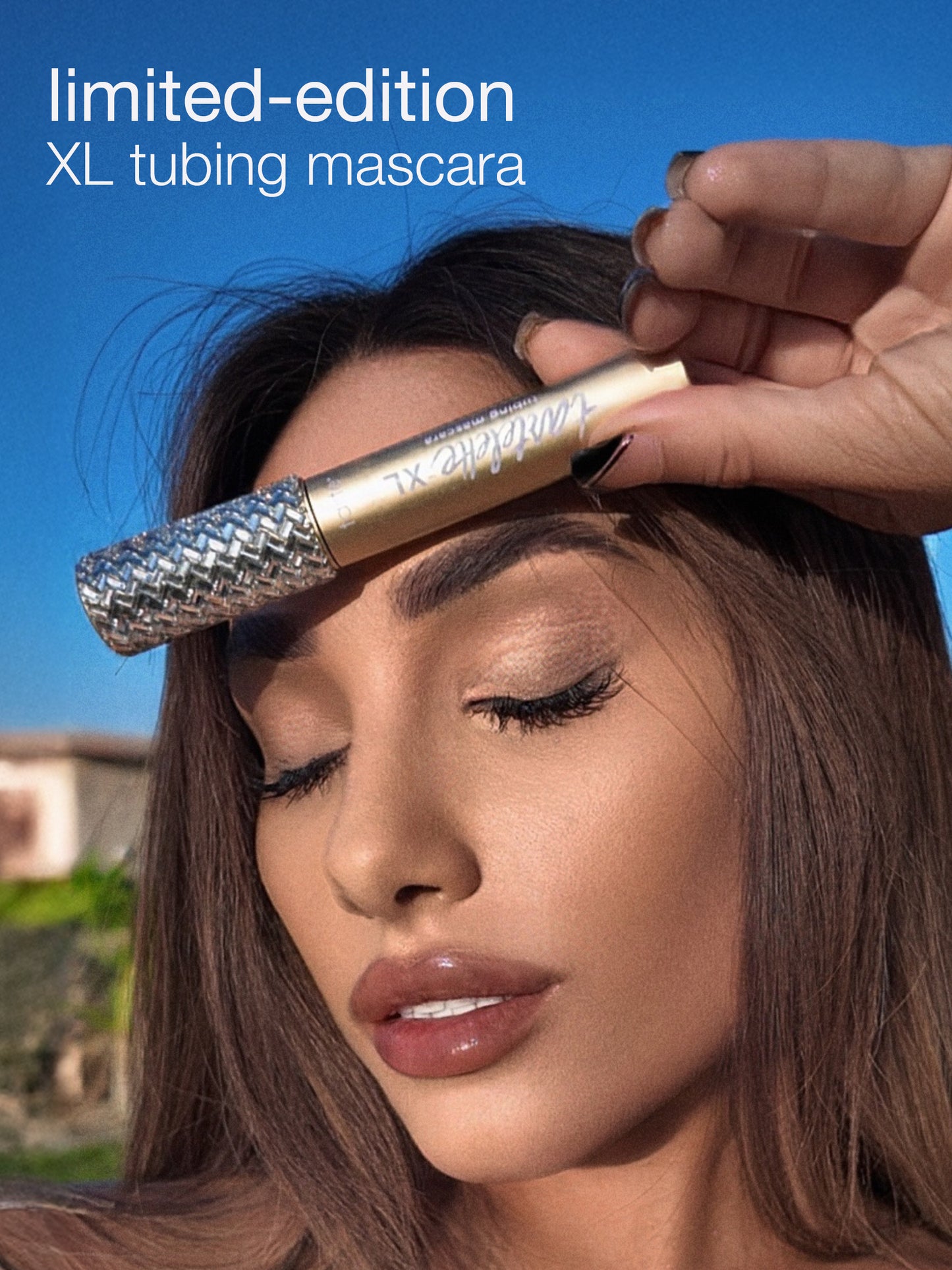 A hand holds a Tarte Cosmetics XL tubing mascara near a woman's closed eyes against a clear blue sky. Text reads, limited-edition XL tubing mascara.