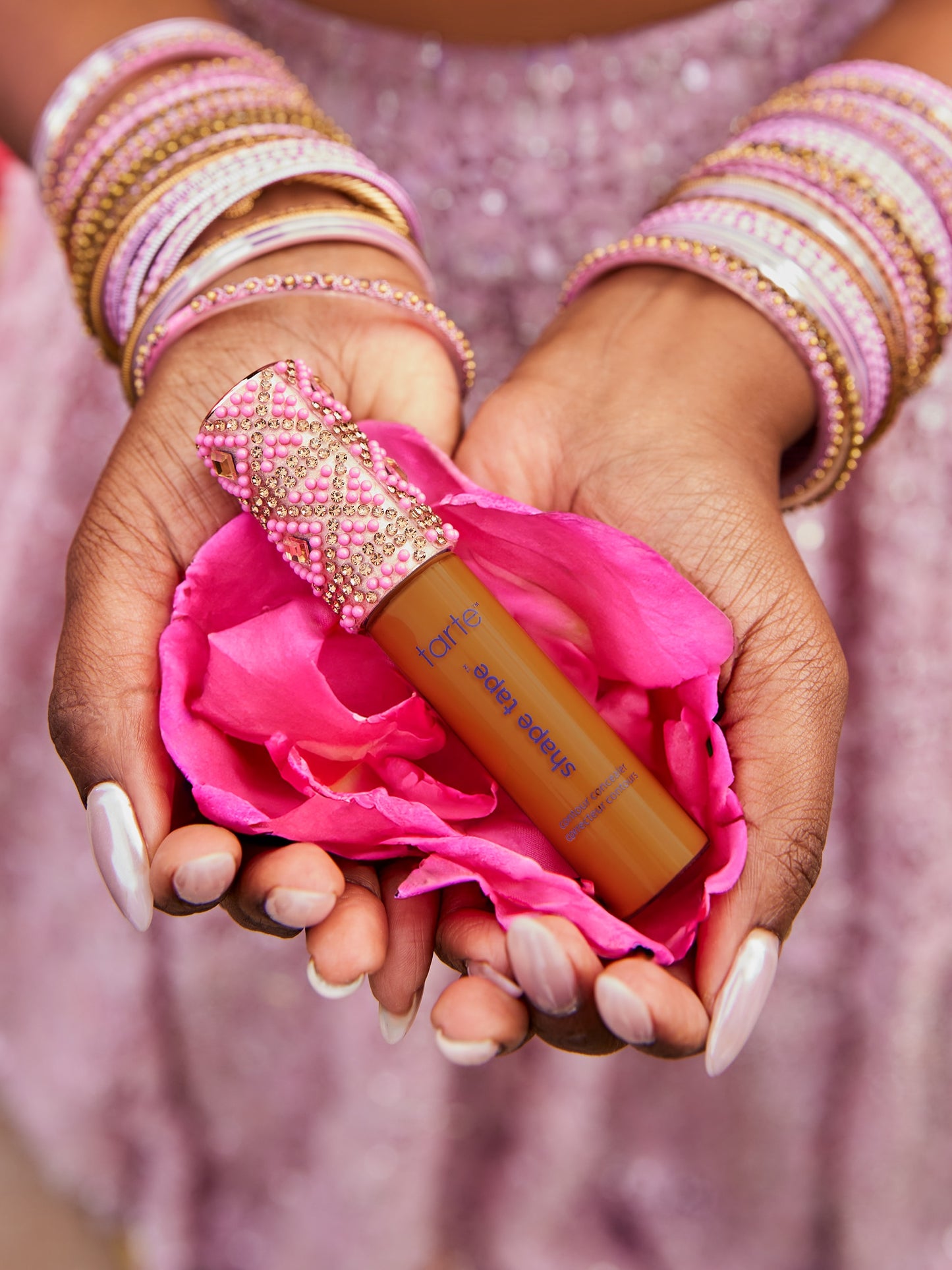 A Tarte Cosmetics Shape Tape concealer rests in cupped hands, adorned with pink and gold bangles, cradled in vibrant pink petals against a sparkling pink fabric background.