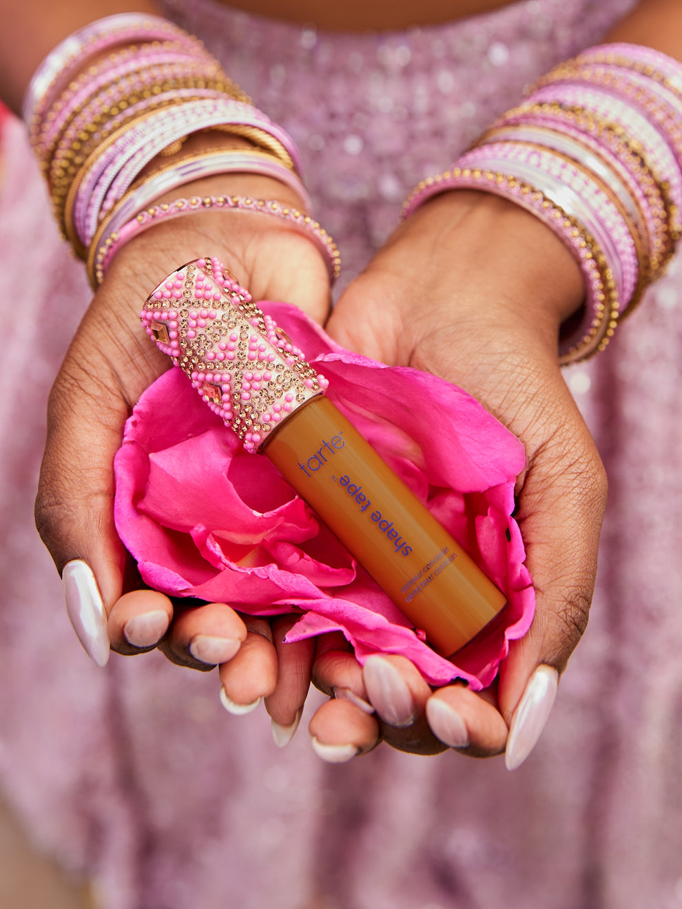 A Tarte Cosmetics Shape Tape concealer rests in cupped hands, adorned with pink and gold bangles, cradled in vibrant pink petals against a sparkling pink fabric background.