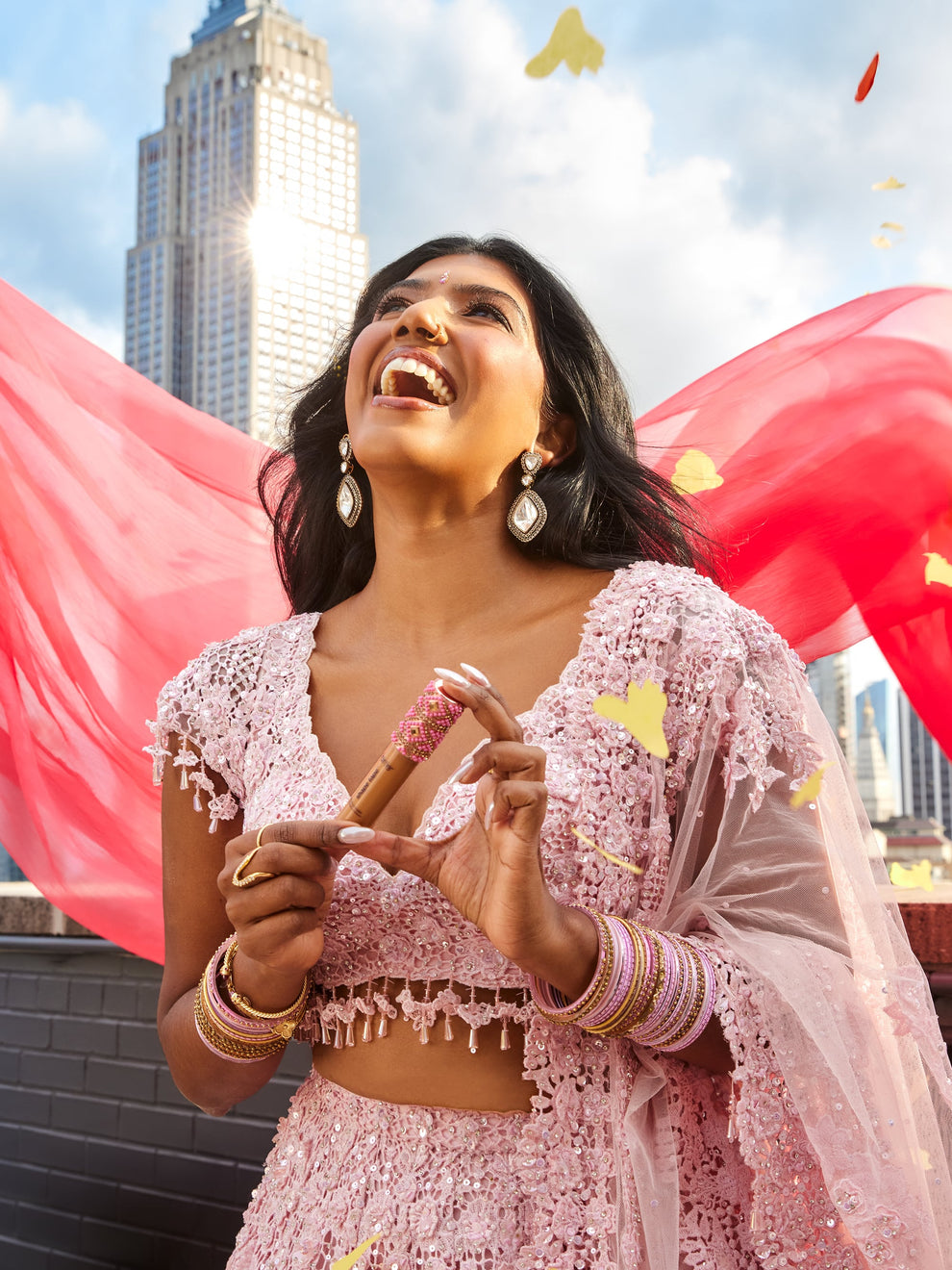 A person in a pink lace outfit joyfully holds a Tarte Cosmetics item amidst fluttering petals, with a cityscape and a tall building in the background.