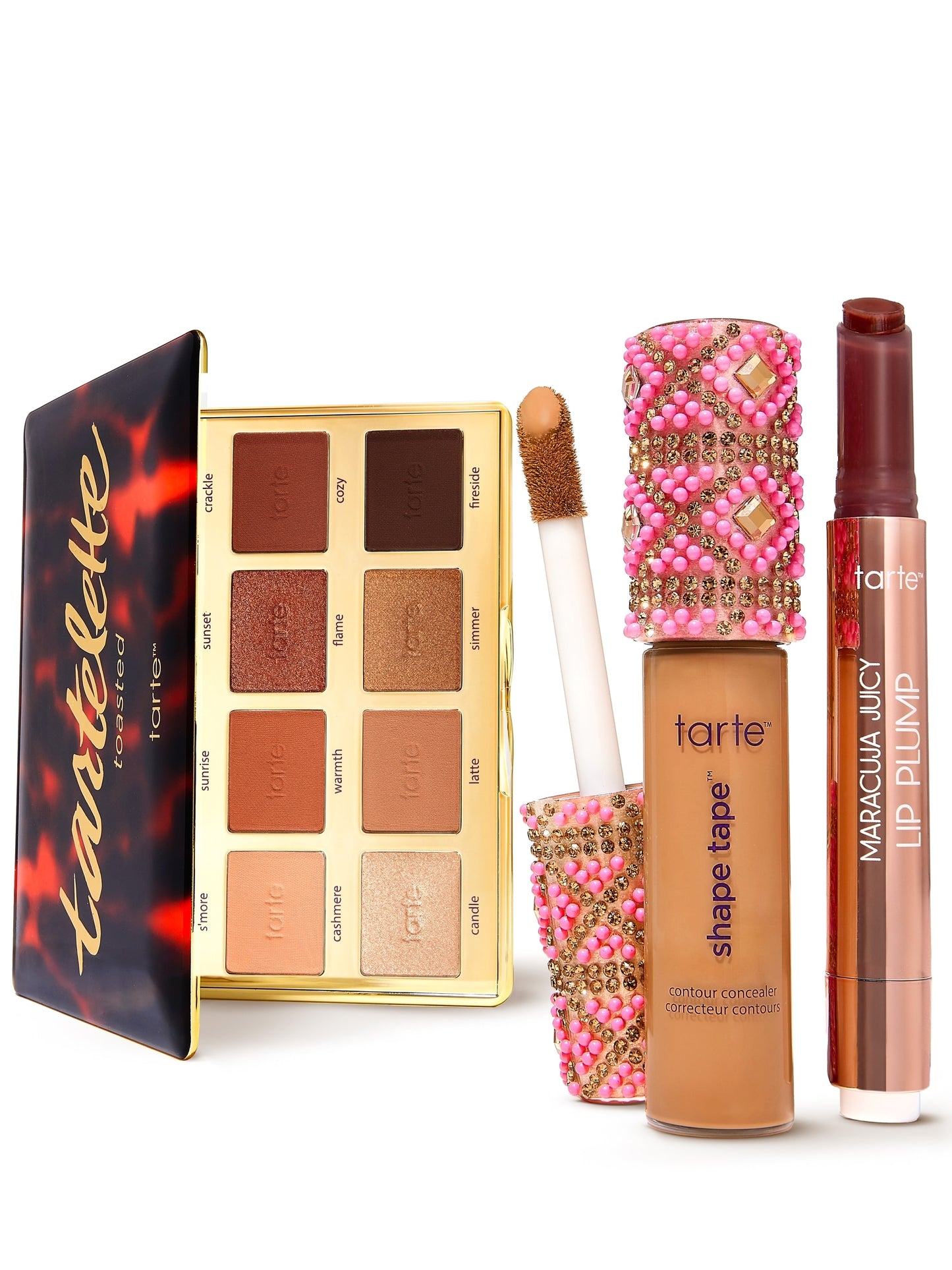 An open eyeshadow palette displays warm shades labeled crackle, cozy, fireside, s'more, sunrise, sunset, warmth, flame, simmer, latte, cashmere, candle. Nearby are Tarte shape tape concealer and Maracuja Juicy Lip Plump, both with decorative elements.