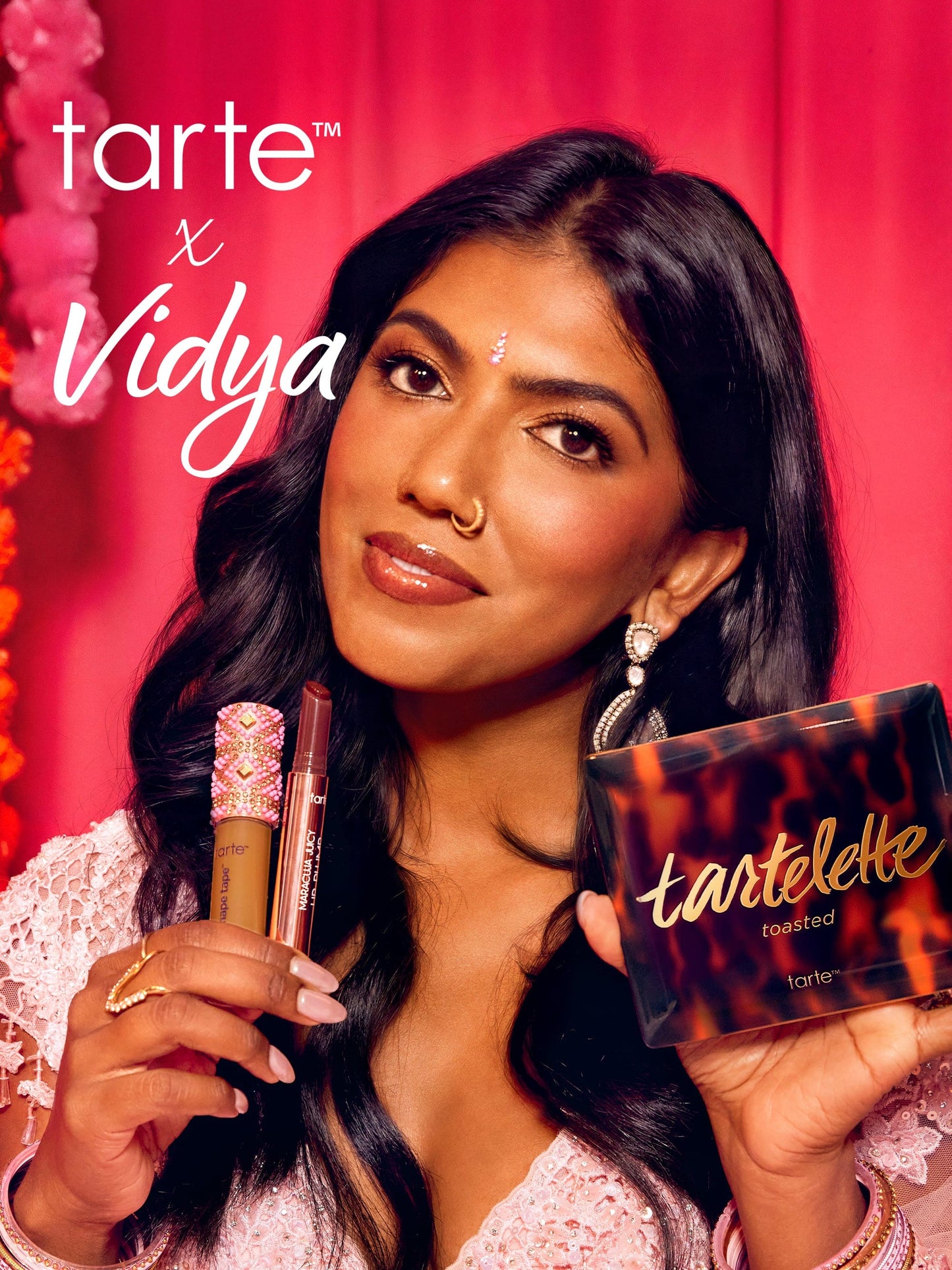 A woman holds Tarte Cosmetics products, including a lipstick and eyeshadow palette, against a vibrant pink backdrop. Text reads tarte™ x Vidya and tartelette toasted. She wears festive attire and jewelry.