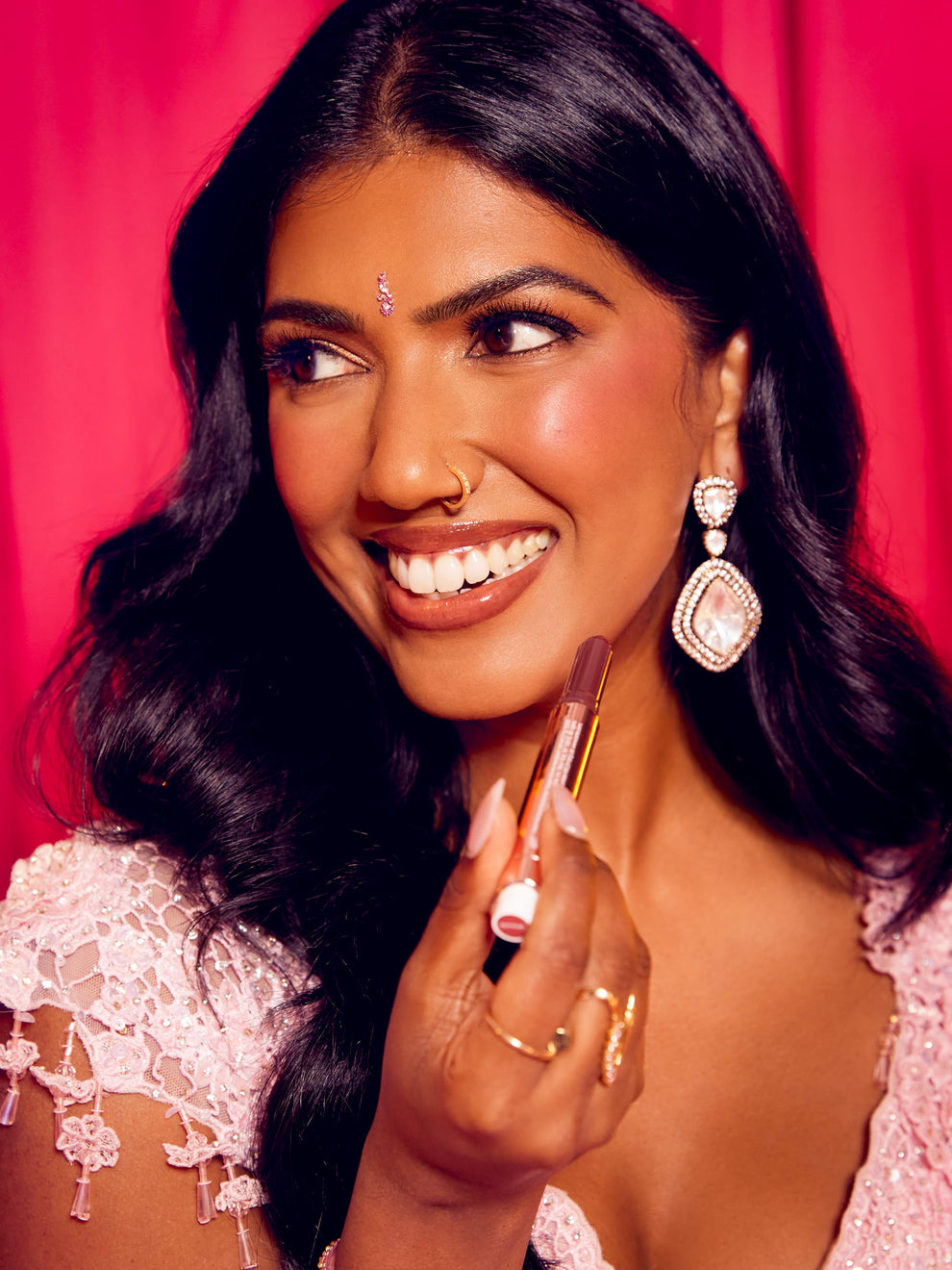 A woman smiles, holding Tarte Cosmetics lipstick near her face. She wears intricate earrings, a nose ring, and a decorative forehead adornment, standing against a pink background.