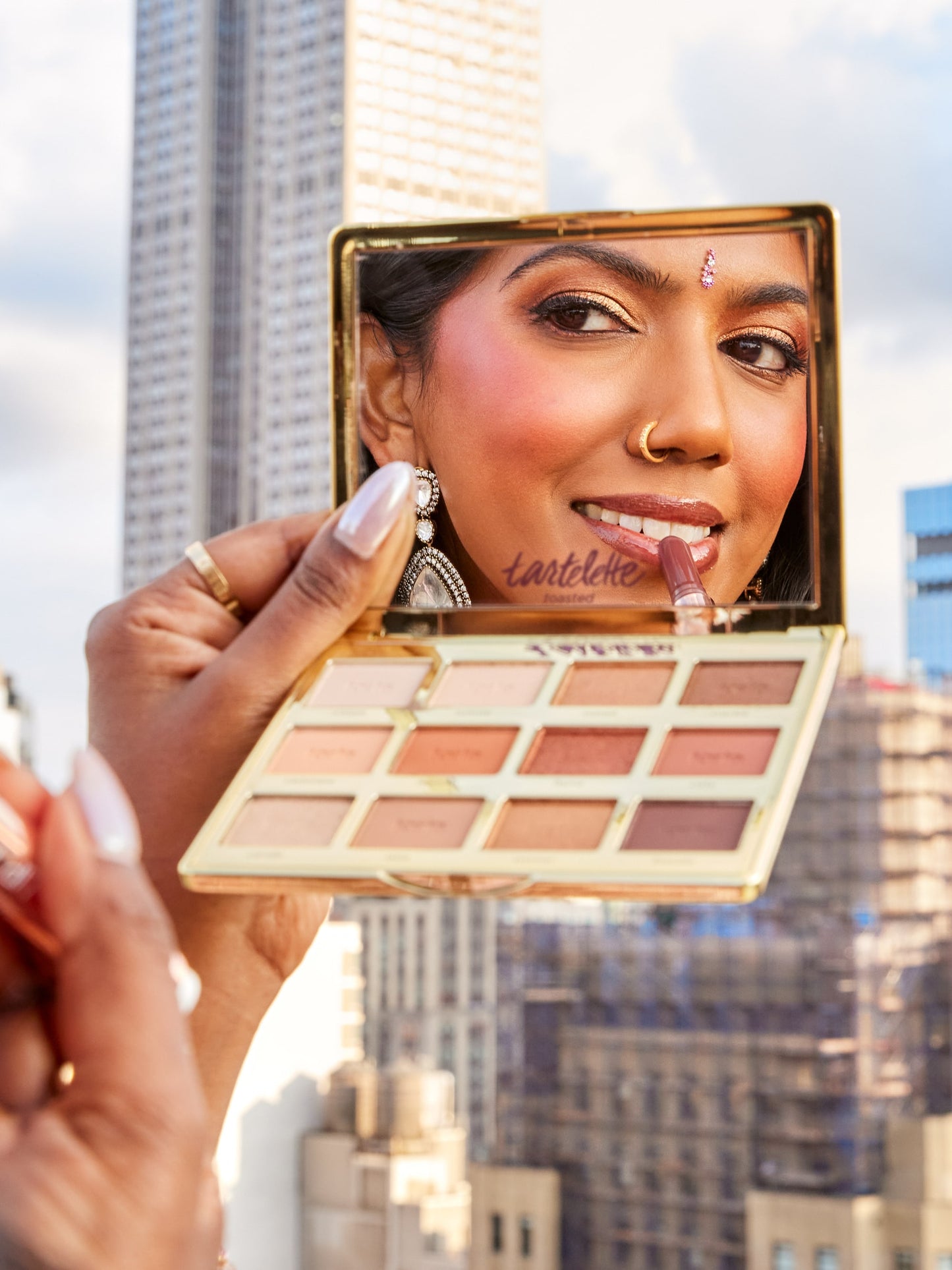 A person applies lipstick while holding a Tarte Cosmetics eye shadow palette mirror, reflecting their face. They're outdoors in a city with tall buildings. The palette reads tartelette toasted.