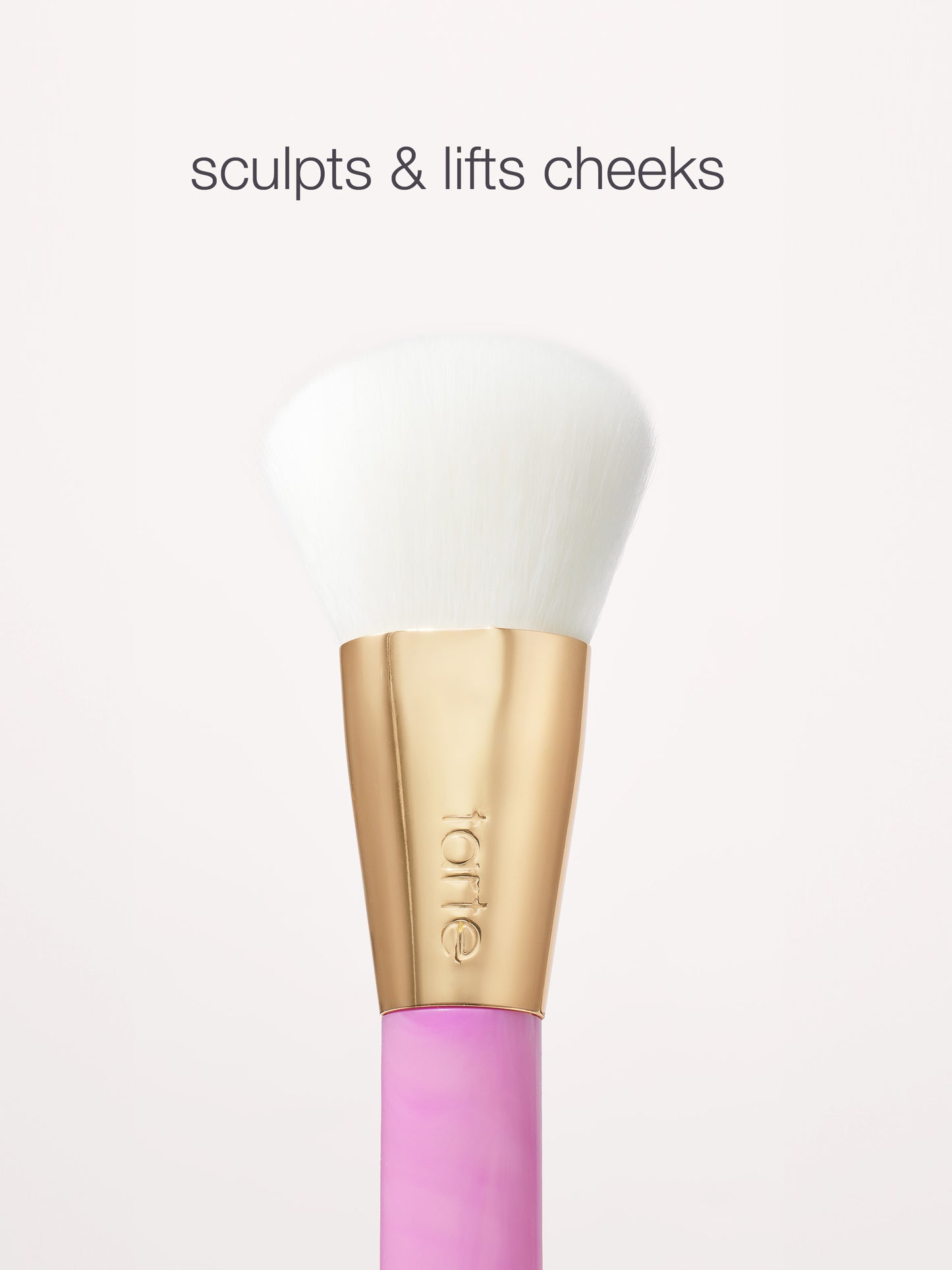 A makeup brush with soft white bristles and a gold handle labeled tarte rests against a light background. Text above reads, sculpts & lifts cheeks.