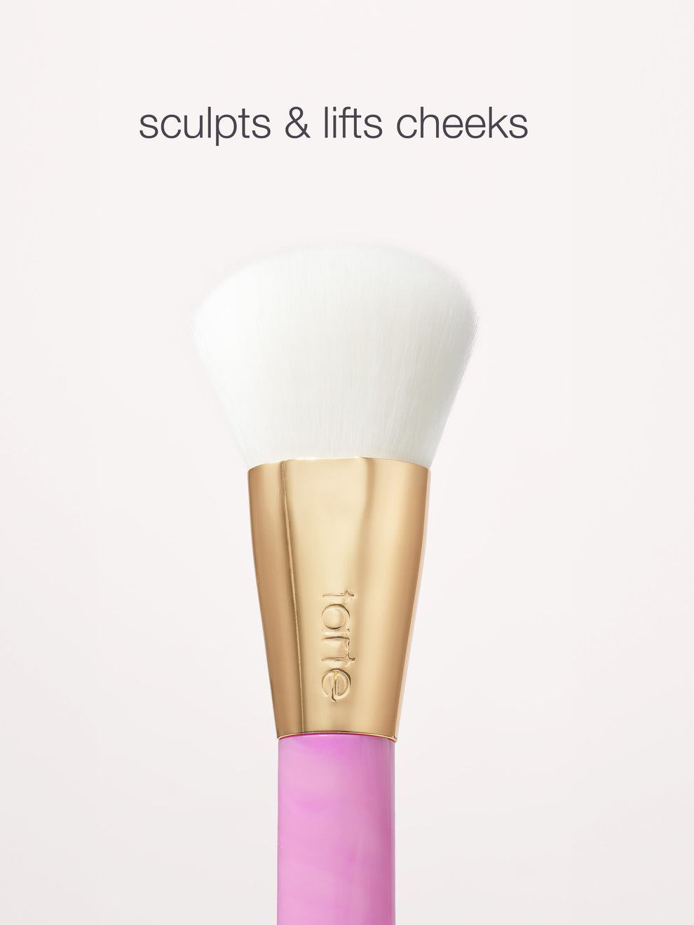 A makeup brush with soft white bristles and a gold handle labeled tarte rests against a light background. Text above reads, sculpts & lifts cheeks.