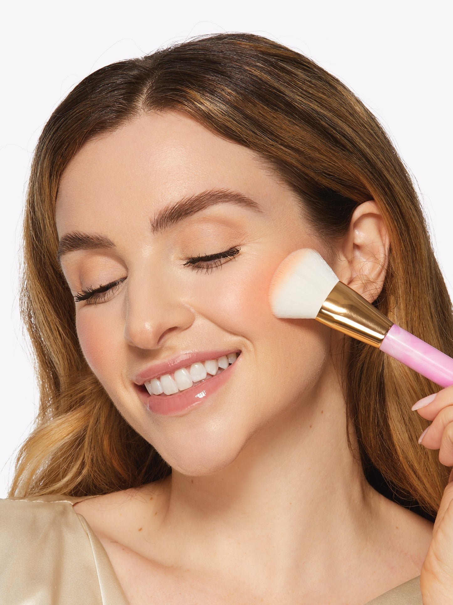 A woman gently applies blush with a Tarte Cosmetics brush to her cheek, eyes closed and smiling, in a well-lit, neutral background setting.