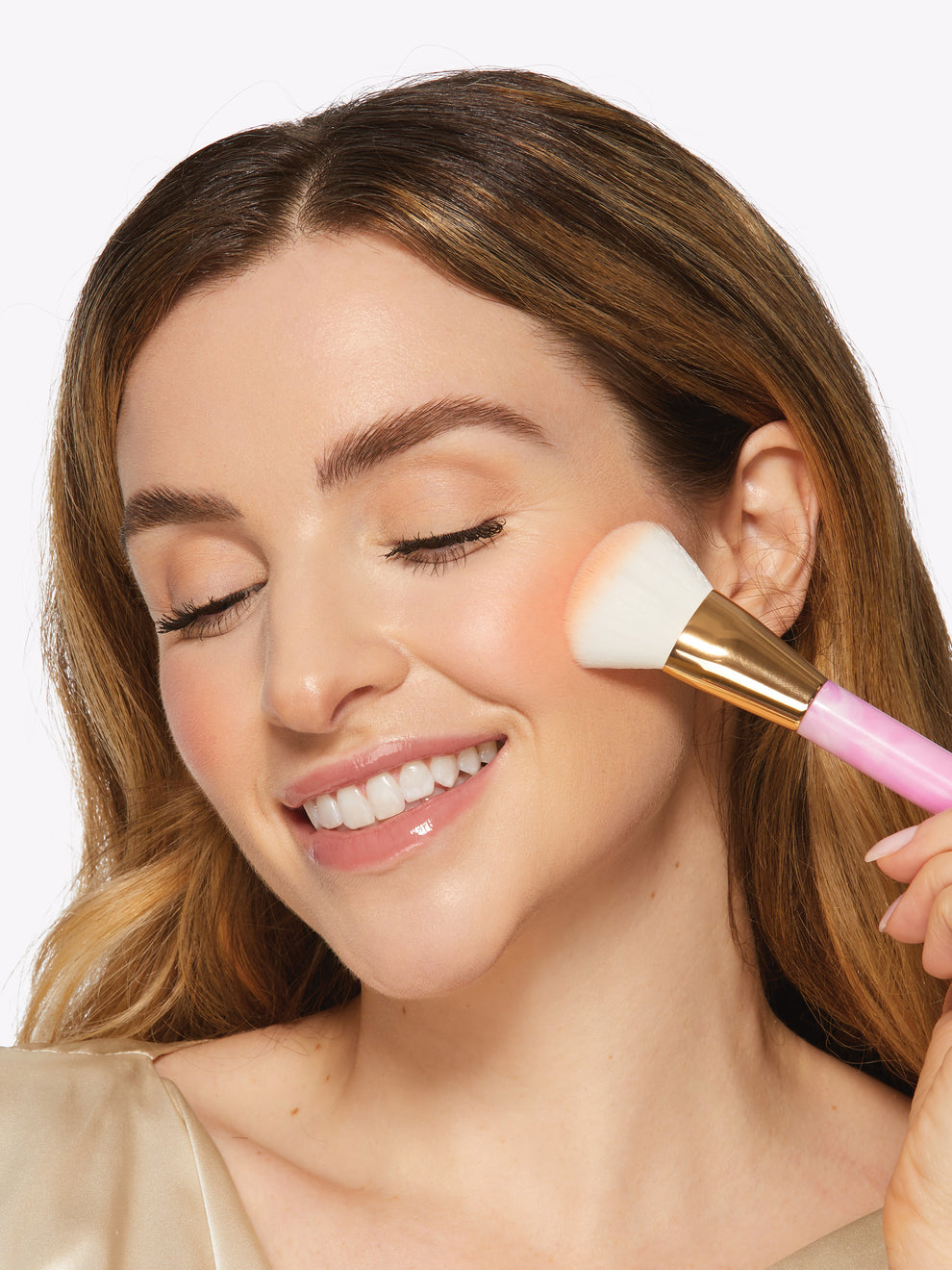 A woman gently applies blush with a Tarte Cosmetics brush to her cheek, eyes closed and smiling, in a well-lit, neutral background setting.