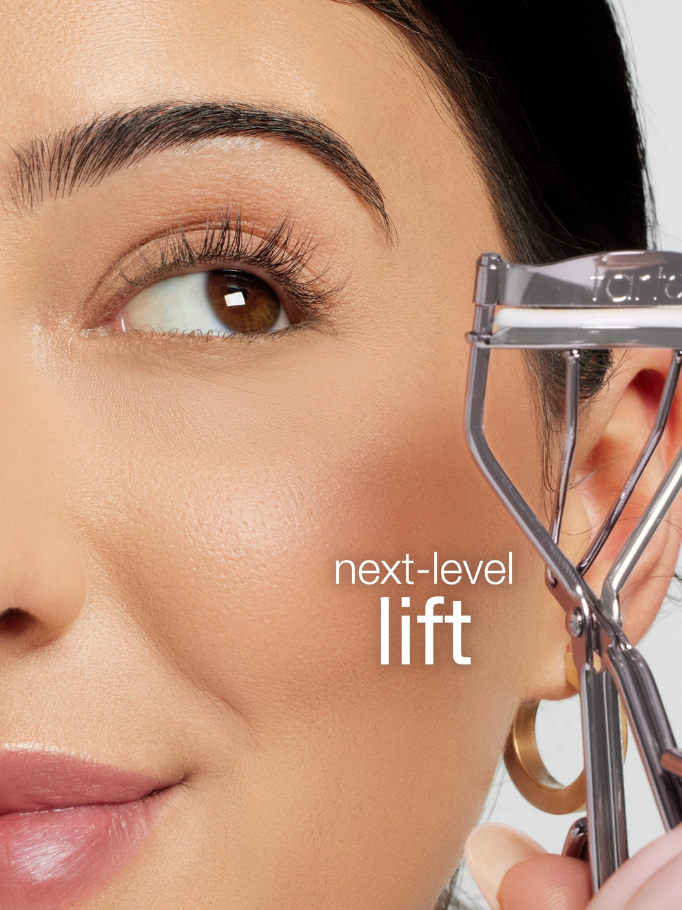 Woman using an eyelash curler with text 'next-level lift' on a blurred background