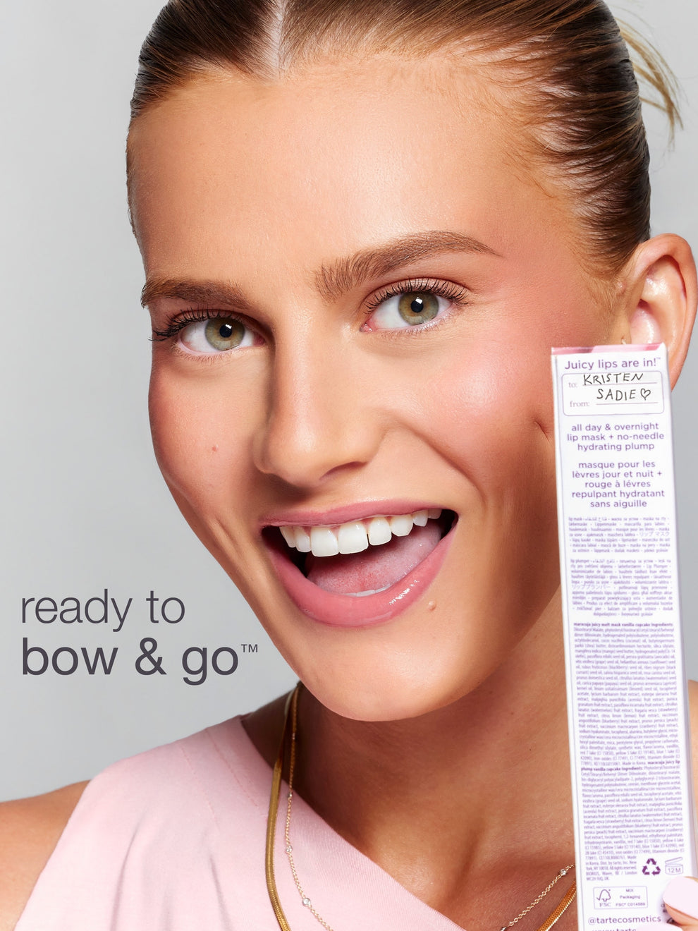 Person smiling brightly, holding up a product package beside their face. Text on the package reads: Juicy lips are in! to: Kristen from: Sadie ♥ all day & overnight lip mask + no-needle hydrating plump. Additional visible text: ready to bow & go™ @tartecosmetics.