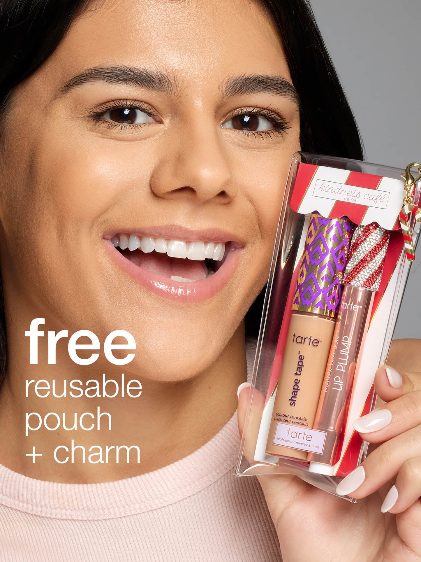 A person smiles while holding a clear pouch containing Tarte Cosmetics products, showcasing a shape tape concealer and Lip Plump. Text reads, free reusable pouch + charm. Sign reads, kindness café est 99.