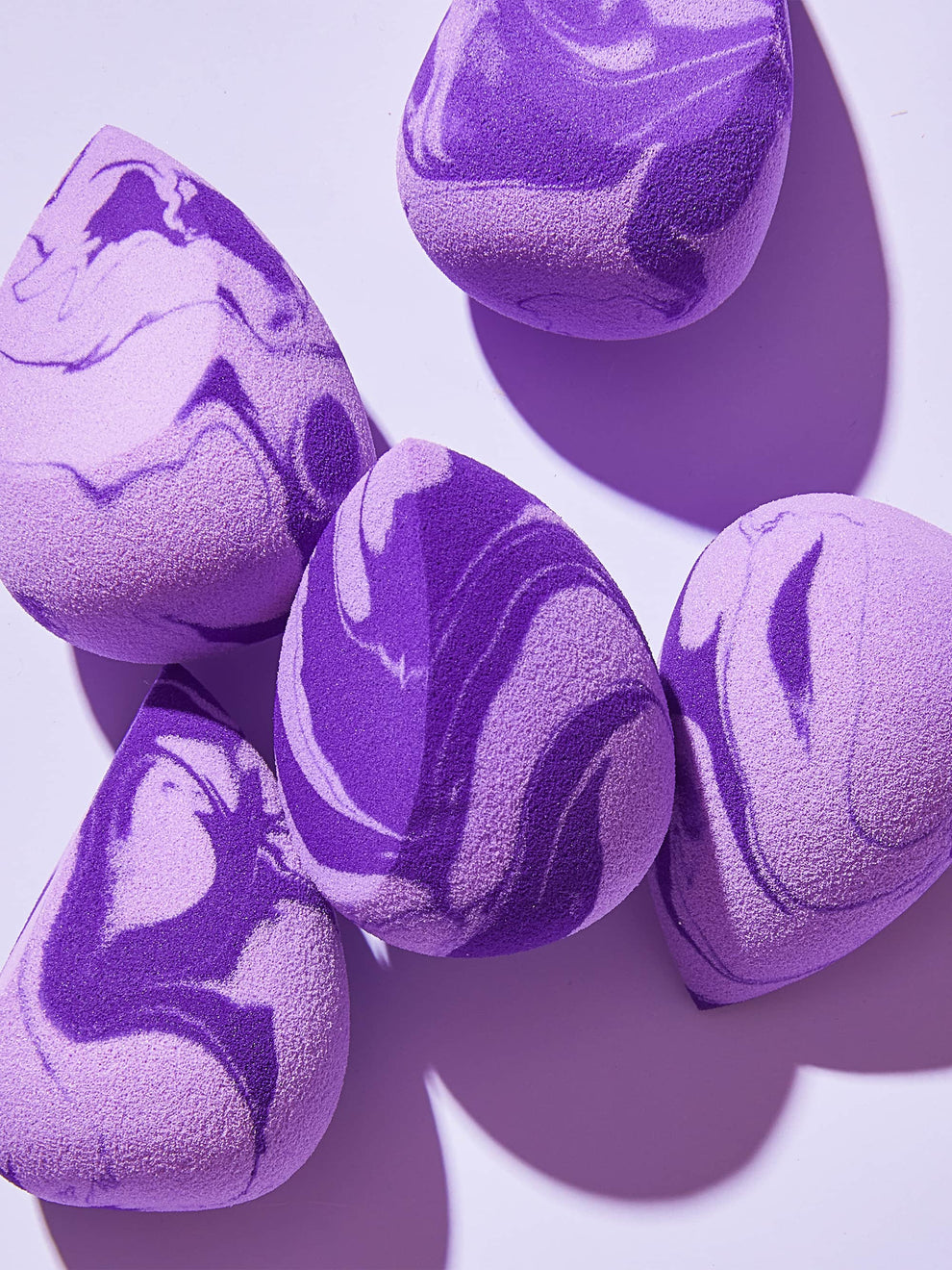 Purple and lavender marbled makeup sponges are scattered on a flat, light-purple surface, casting soft shadows in a minimalist setting.