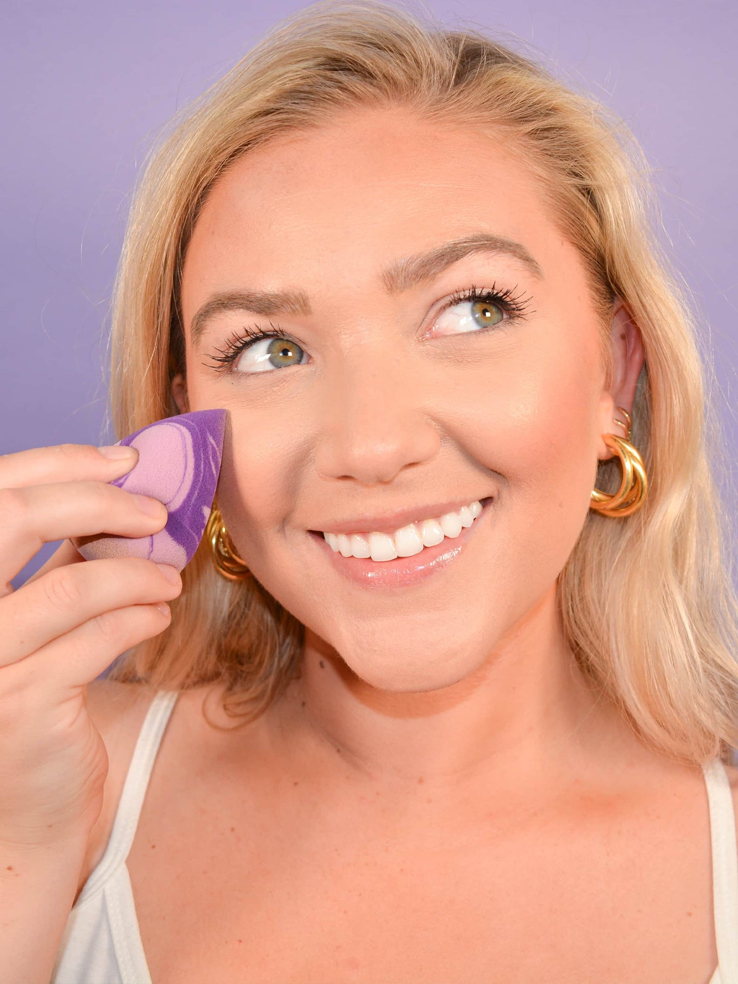 A person applies makeup with a purple sponge while smiling, wearing gold hoop earrings. The background is a soft purple, enhancing the warm and cheerful ambiance.