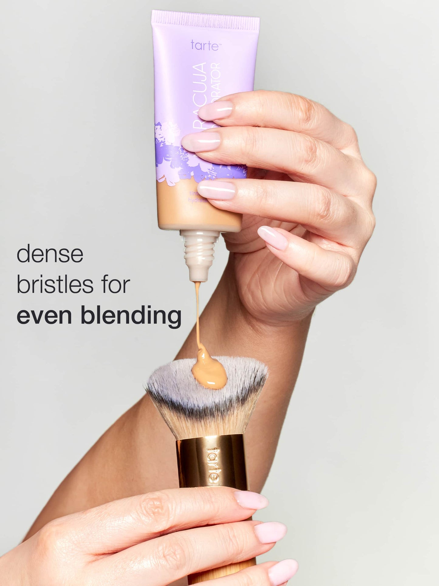 Hand holding a makeup brush with foundation and a Tarte product on a gray background