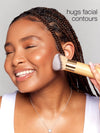 Person applying makeup with a brush to their face, with text 'hugs facial contours' on a gray background.