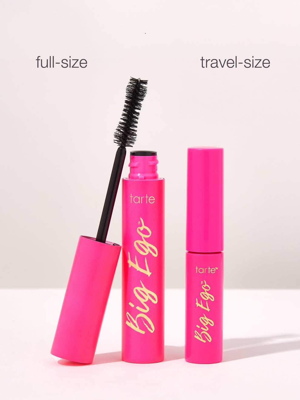 A full-size and travel-size pink tube of Tarte Big Ego mascara stand upright. The full-size tube is open with a brush extended. The words full-size and travel-size are labeled above.
