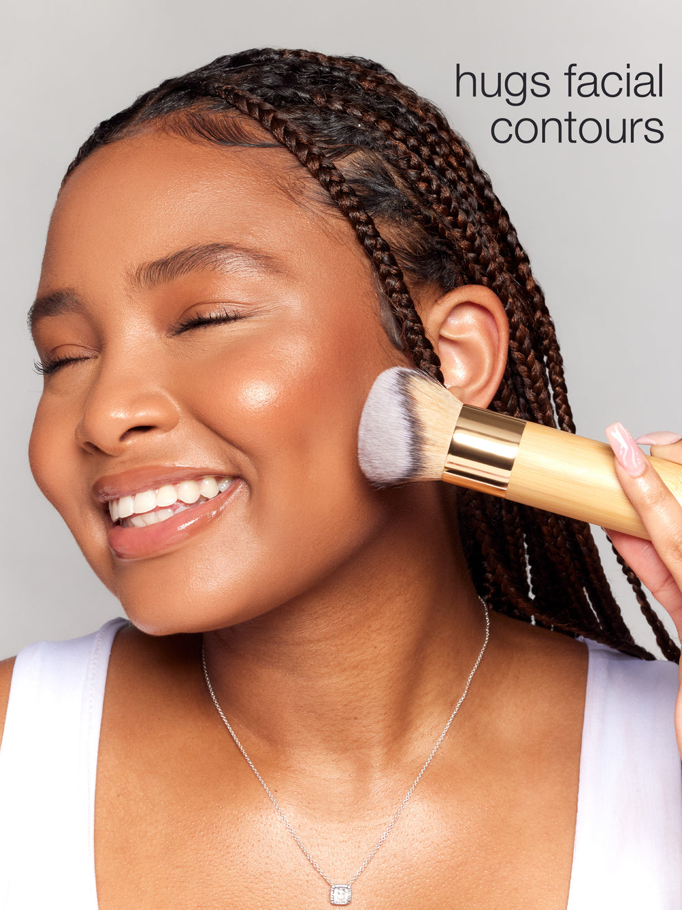 Woman applying makeup with a brush to her face, with text 'hugs facial contours' on a gray background.