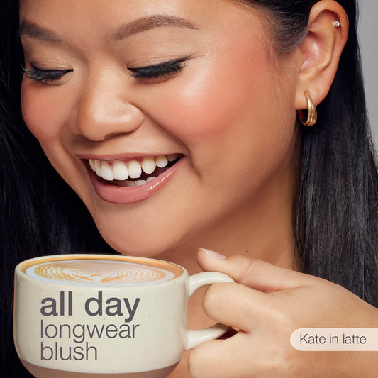 A smiling person holds a latte cup, showcasing rosy cheeks and gold hoop earrings. Text on the cup reads: all day longwear blush. Additional text says: Kate in latte.