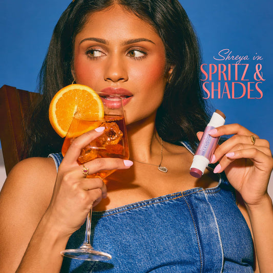 Woman holding a glass of orange juice with an orange slice and a blush, bronzer stick, against a blue background with 'Spritz & Shades' text.