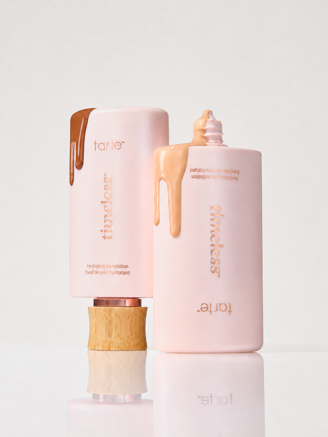 Two Tarte Timeless foundation bottles in a soft blush pink color with gold lettering and decorative cream-colored and brown cream drips on the caps, displayed on a reflective white surface against a light gray background.