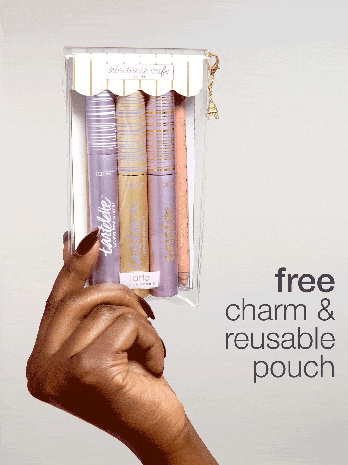 A hand holds a transparent pouch containing Tarte Cosmetics products, adorned with a decorative charm. The text on the pouch reads kindness café est 99. To the right, bold text states free charm & reusable pouch.