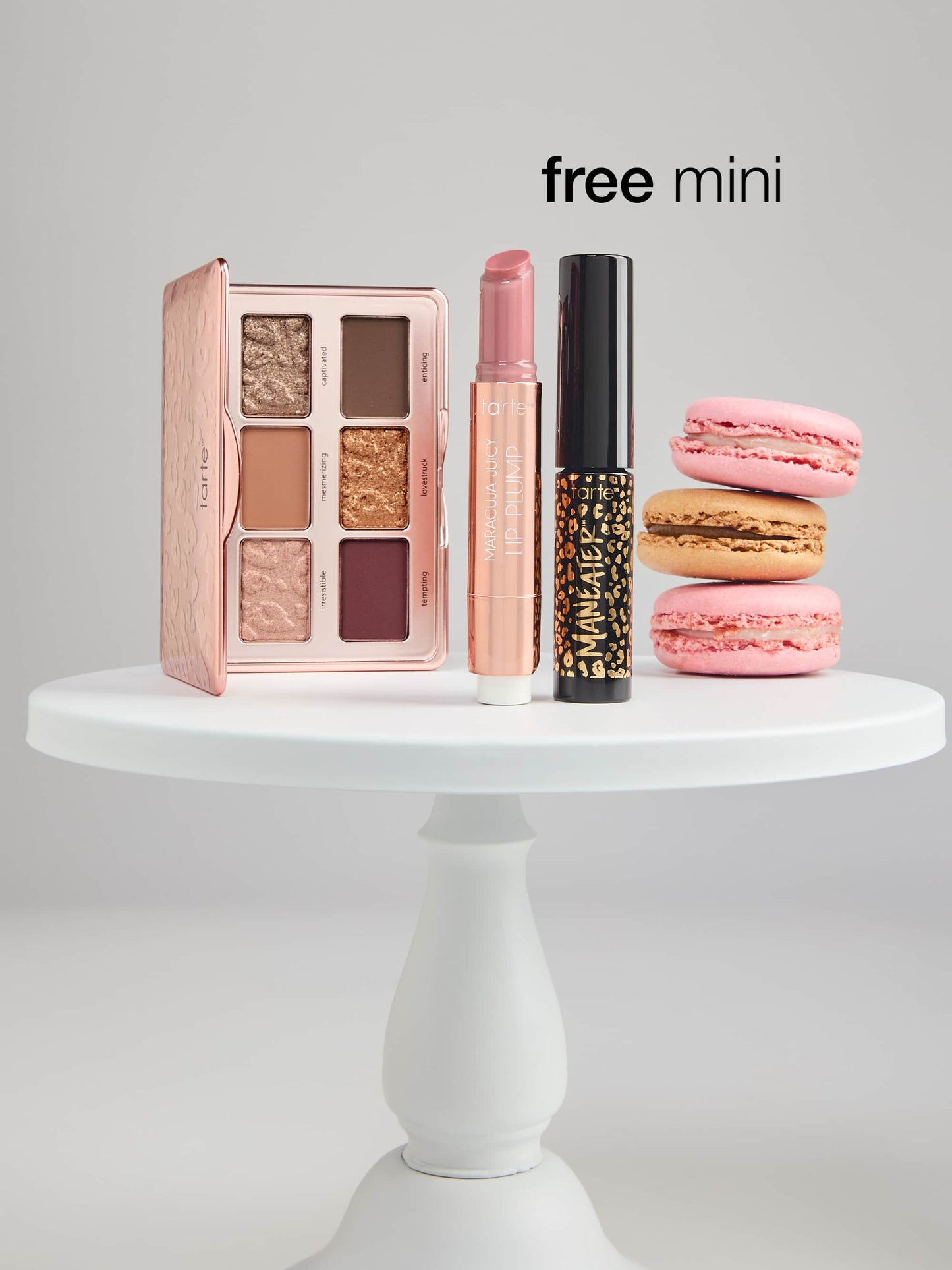 Makeup products including an eyeshadow palette, lip plump, and mascara are displayed on a white pedestal with macarons nearby. Text reads free mini.