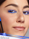 Close-up of a woman's face with blue eyeliner and mascara, with a blue mascara wand in the foreground.