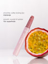 A lip gloss tube leans against a halved passion fruit on a white surface. Text reads: smoother, softer-looking lips maracuja smooth, nourish & hydrate TIA superfruits.