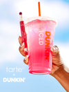 Hand holding a pink iced drink with an orange straw and a red lip product, against a blue gradient background. 'tarte' x 'Dunkin'' branding visible.